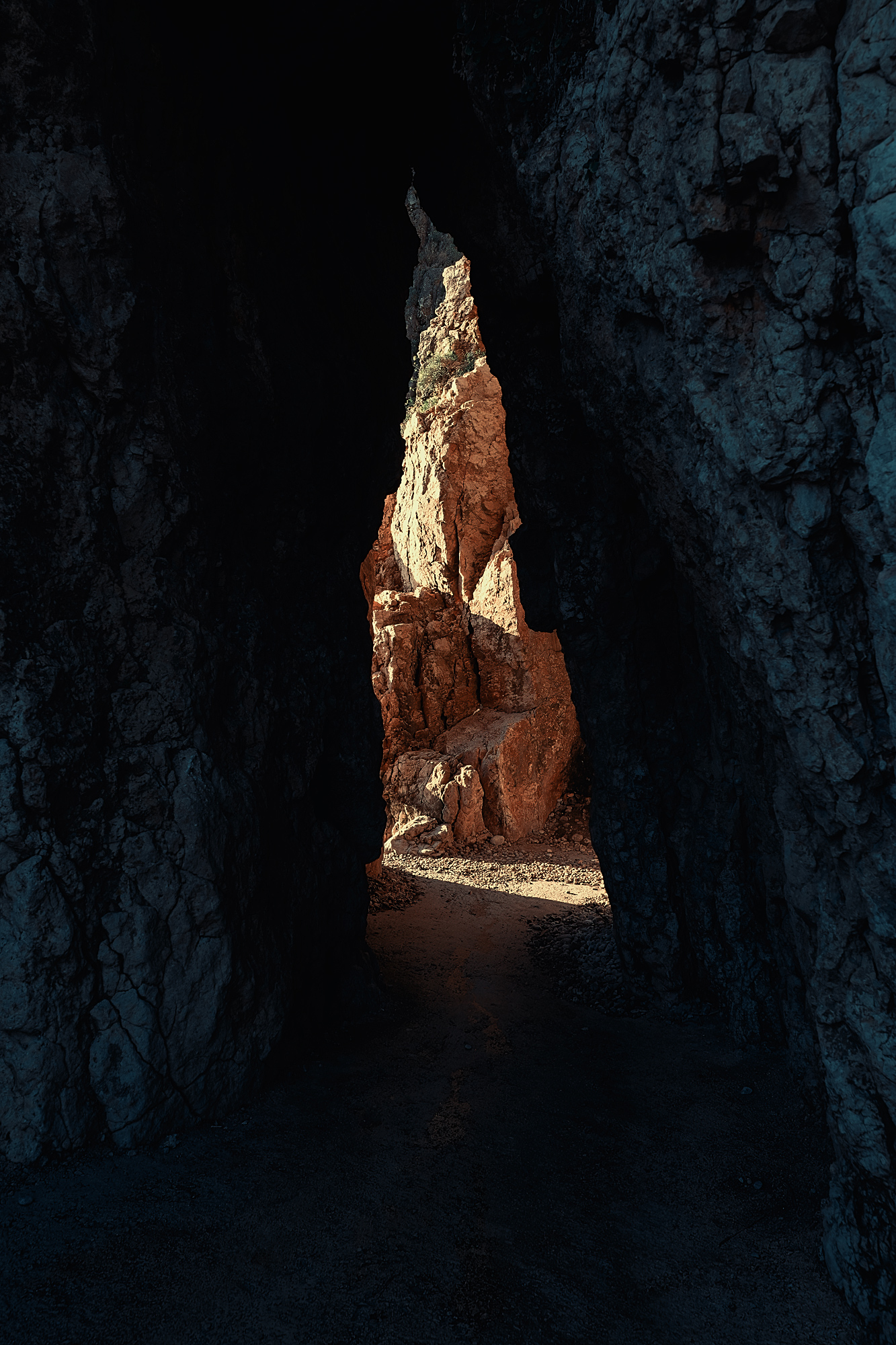 A narrow gap between dark rock walls frames a sunlit orange rock face and a strip of pebble-covered ground.