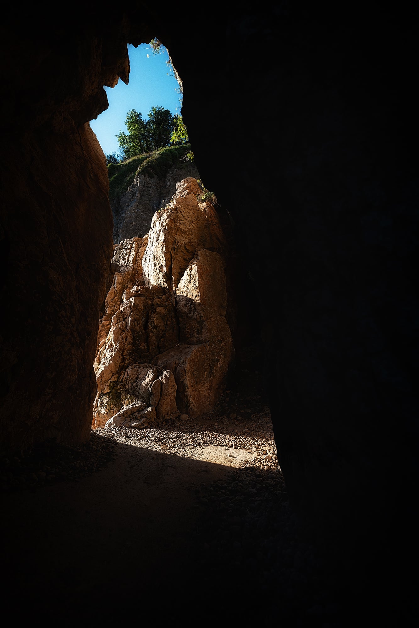 Dark overhanging rock frames a sunlit formation, pebbles, green shrubs and a narrow opening to blue sky.