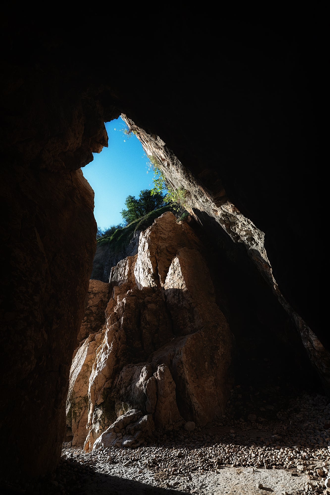 Blue sky and sunlit orange rock seen through an angular opening above a pebble-covered floor.