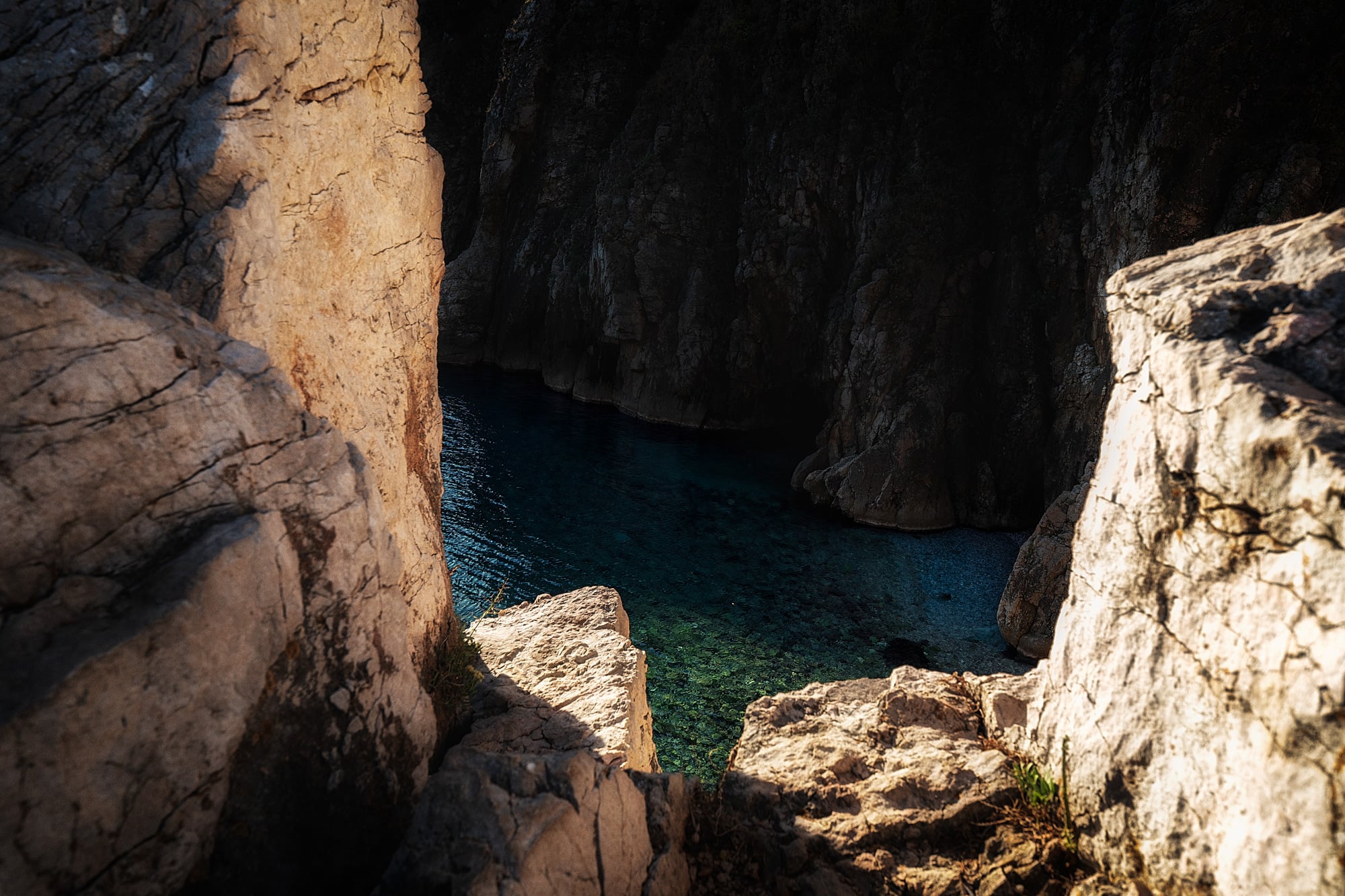 Clear blue-green seawater lies between dark cliffs and pale sunlit rocks in the foreground.