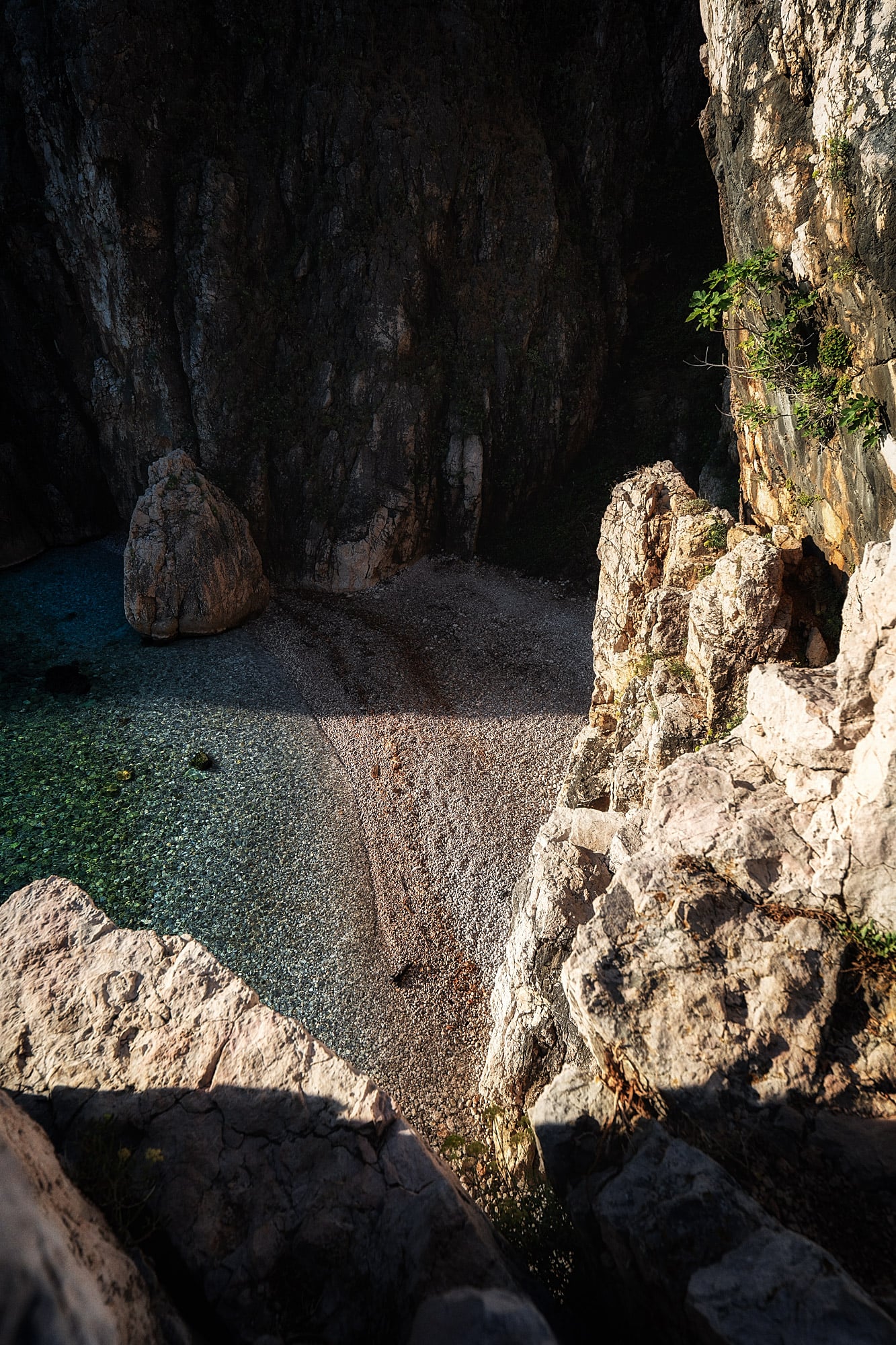 A small pebble beach curves beneath steep shadowed cliffs beside clear water and a large boulder.