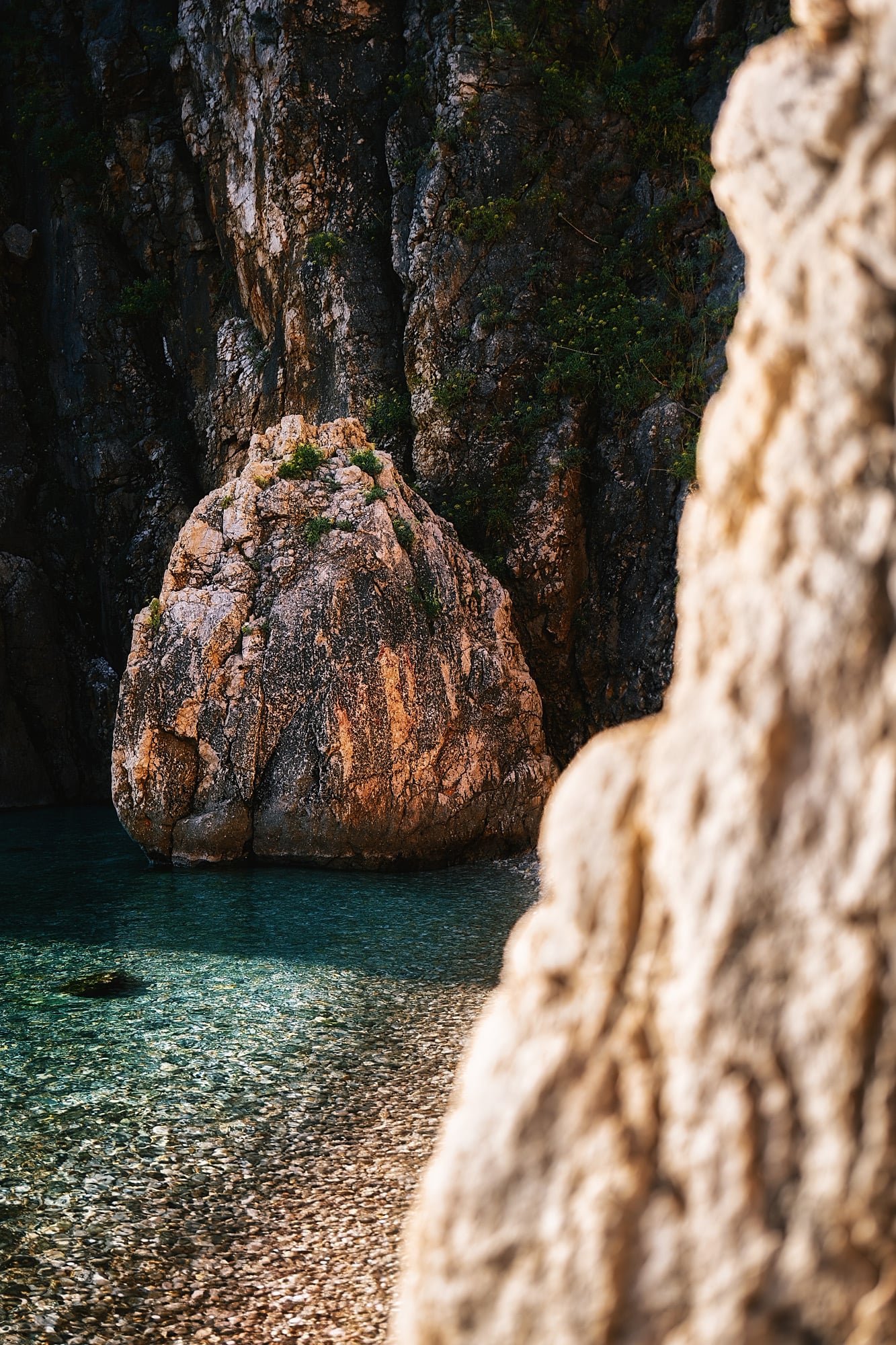 A rounded orange-gray boulder stands in clear shallow water, framed by an out-of-focus rock on the right.