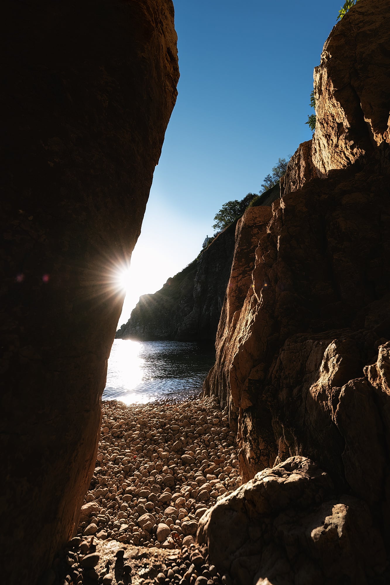 Sunlight bursts around a dark rock edge above a pebble shore, with blue sea and sunlit cliffs beyond.